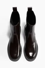Botas Chelsea de cuero sintético color marrón oscuro (casi negro), con elásticos laterales negros y trabilla trasera. Tienen suela de goma negra con relieve y un taco bajo y ancho.