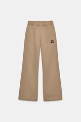 Pantalón de felpa color beige, tiro medio, con cintura elástica, bolsillos laterales ocultos y pierna ancha. Presenta un parche pequeño con un logo en la parte delantera.