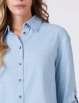 Camisa de algodón y lyocell, marca Weekend Routine, color celeste con finas rayas blancas. Tiene cuello con solapa, botones metálicos en la abertura frontal y mangas largas regulables con presilla y botón. La espalda presenta un canesú horizontal con un tablón central, y el bajo es asimétrico y redondeado.