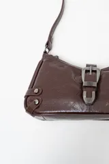 Bolso de hombro tipo baguette en color negro con acabado brillante. Presenta un diseño estructurado con una hebilla decorativa metálica en el frente y detalles de tachas plateadas en los laterales.