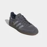 Championes Adidas Handball Spezial de gamuza color gris con las icónicas tres tiras laterales en un tono más claro y suela de caucho natural. Presenta el logo de la marca y el nombre del modelo en dorado en el lateral.