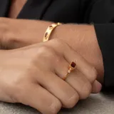 Anillo dorado con piedra central rectangular color borgoña y dos diamantes circulares a cada lado.