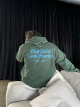 Canguro verde oliva con capucha y cordones ajustables. Estampado en la espalda con la frase "Bad Ideas Good Stories" en letras azules.
