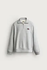 Sudadera gris de corte relaxed fit con cuello alto y cierre frontal de cremallera corta, confeccionada en algodón con interior de rizo afelpado. Presenta mangas largas, puños y bajo de rib, y un parche bordado con el logotipo de Fruit of the Loom en el pecho. Colección especial Fruit of the Loom x Zara.