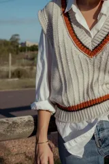 Chaleco tejido color crema con cuello en V y ribetes en naranja y negro. Diseño sin mangas y corte holgado. Ideal para usar sobre camisas o remeras de manga larga.
