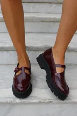 Zapato tipo guillermina en cuero charol color bordó, con suela track negra de goma, cierre con hebilla ajustable y detalle de herrajes metálicos dorados.