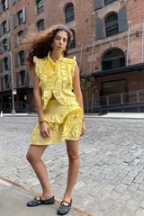 Blusa amarilla sin mangas, con cuello alto y cierre frontal abotonado. Presenta abundantes volados decorativos a lo largo de la abertura y en los hombros.