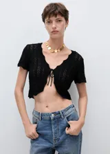 Cardigan de punto calado color negro, con mangas cortas, cuello en pico y cierre frontal mediante lazo fino. Diseño corto y ajustado.