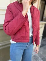 Campera tipo bomber acolchada con diseño de pespuntes ondulados, cierre frontal y bolsillos laterales con botón. Presenta un corte moderno y acabado en color bordo.