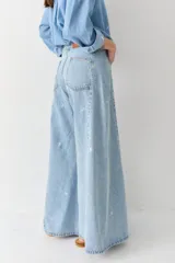 Jean de corte wide leg y tiro alto, confeccionado en denim de lavado claro. Presenta un diseño con costuras verticales frontales y un sutil estampado de figuras deportivas pequeñas distribuidas por toda la prenda.