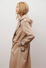 Trench coat color beige con cuello de solapa, cierre frontal abotonado, bolsillos laterales y mangas largas con cintas ajustables. Incluye capucha desmontable y forro interior rayado. La tela exterior cuenta con protección repelente a la lluvia y manchas.
