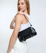 Cartera tipo baguette de tamaño pequeño, confeccionada en poliuretano color negro. Presenta una solapa frontal con detalles de hebillas metálicas decorativas en los laterales y un asa de hombro ajustable.