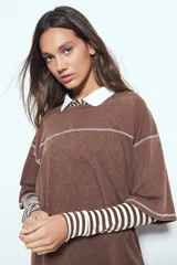 Remera de manga corta color marrón con efecto desgastado, corte oversize y costuras a la vista en color blanco. Presenta cuello redondo con cuello polo blanco superpuesto y mangas largas a rayas horizontales marrones y blancas por debajo.