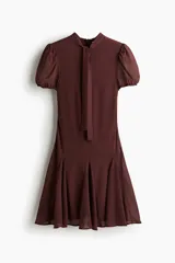 Vestido corto color marrón oscuro, con cuello alto y lazo frontal. Presenta mangas cortas abullonadas y una falda con vuelo y pliegues.