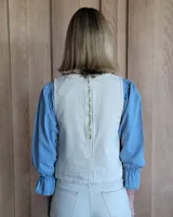 Chaleco de corte recto y largo a la cintura, confeccionado en denim con estampado de rayas verticales en tonos azul y blanco. Presenta cuello redondo y costuras visibles que definen su estructura.