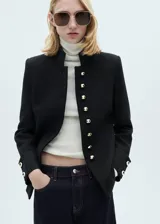 Chaqueta estructurada de corte recto en color negro, con cuello mao y cierre frontal de botones dorados con efecto joya. Presenta manga larga con puños abotonados y forro interior.