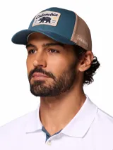 Gorro trucker Columbia de color verde azulado y beige, con paneles de malla trasera y un parche frontal estampado con un oso y la marca.
