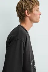 Camiseta de punto con corte boxy fit, cuello redondo y manga corta. Color negro con efecto lavado y terminaciones irregulares. Presenta un estampado frontal con texto y un gráfico circular en tonos blanco y rojo.