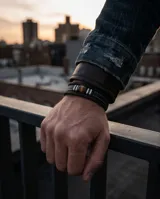 Pulsera de diseño masculino compuesta por bandas de cuero sintético trenzado en color negro, con un elemento central de piedra ojo de tigre flanqueado por detalles metálicos de acero.