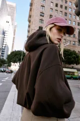 Chaqueta estilo bomber de corte oversize en color marrón oscuro, confeccionada en tejido suave. Presenta capucha con cordones ajustables, cierre frontal con cremallera, mangas largas con puños elásticos y bolsillos delanteros de vivo. El bajo cuenta con detalle de pliegues.