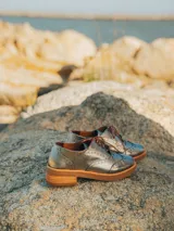 Zapato acordonado estilo oxford de cuero vacuno en color plateado metalizado, con diseño brogue, suela de goma y taco de madera.