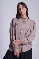 Camisa de color azul con cuello alto decorado con volados y lazo de ajuste. Presenta tablas decorativas en los hombros, cierre frontal con botones de nácar y mangas largas con puños elásticos que generan un efecto abullonado.