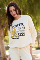Sweater de tejido de punto color crema, con cuello redondo y manga larga. Estampado frontal con botas de cowboy amarillas y texto que dice "Boots Class and Little Sass".