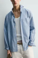 Camisa de hombre de manga larga, corte relajado (relaxed fit), color celeste con patrón de cuadros pequeños en azul y rojo. Presenta cuello abotonado (button-down), bolsillo de parche en el pecho y cierre frontal de botones.