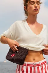 Mini bolso de mano tejido en rafia color marrón oscuro, con diseño estructurado y detalle de flor decorativa en tonos rojizos realizada en crochet.