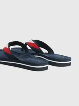 Ojotas de mujer color azul marino con tira de tela trenzada que presenta franjas rojas y blancas, y un dije metálico con el monograma TH en dorado.