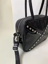 Cartera rígida negra con detalles en rojo y tachas plateadas. Tiene herrajes y cierre metálico plateado marca YKK, asa fina para llevar al hombro o en la mano y correa larga extra regulable. El interior es forrado, tiene un bolsillo con cierre enmarcado en cuero, y un segundo bolsillo plaqué.