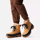 Bota de invierno Sorel modelo Tivoli V, confeccionada en gamuza color tostado con base de goma negra impermeable. Presenta cuello con forro de piel sintética, cordones negros y suela dentada de goma en color marrón.