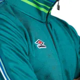 Campera negra de poliéster con cierre de cremallera y cuello alto. Presenta puños y cintura acanalados con rayas grises y blancas. Logo de Umbro bordado en el pecho.