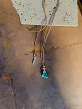 Collar con cordón de cuero color beige y dije de piedra turquesa irregular con detalles plateados. Incluye un dije rectangular plateado con la palabra 'STRONG'.