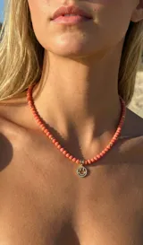 Collar de cuentas de cristal facetado en color coral, con pequeños separadores dorados y un dije colgante en forma de cara feliz (smiley) con apliques de circón, en acero con baño dorado.