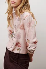 Camisa de manga larga con cuello redondo y cierre frontal de botones. Presenta un estampado floral en tonos azules y marrones sobre una base de color rosado claro.