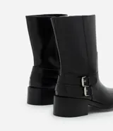 Bota de caña corta marca Satinato, confeccionada en poliuretano color negro. Presenta punta redondeada, taco bloque bajo y dos tiras decorativas con hebillas metálicas en el lateral externo.