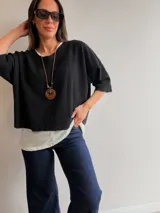 Conjunto de dos piezas compuesto por una remera básica blanca de algodón y un buzo liviano de textura plisada en color azul grisáceo. El buzo presenta un diseño de manga murciélago, corte holgado y detalles de hilo plateado en los bordes. Incluye un collar largo con dije circular de madera y metal.