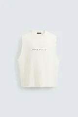 Camiseta sin mangas (manga sisa) de punto color crema, corte relaxed fit, con cuello redondo y acabados irregulares en las sisas. Presenta un texto estampado en el pecho que dice "SERIAL N. 8763/ 7.3Z".