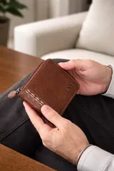 Billetera de cuero marrón oscuro con cierre perimetral y detalle de costura artesanal en el frente. Cuenta con compartimentos internos para billetes, tarjetas y monedero.