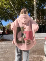 Campera tipo hoodie oversize de corderito rosa, con diseño de collage en la espalda que combina parches de tela en tonos verde oliva, rosa y estampados geométricos étnicos.