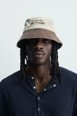 Sombrero bucket confeccionado en tejido de algodón con efecto lavado, diseño bicolor en tonos beige claro y marrón oscuro. Presenta ala ancha con pespuntes y un bordado frontal en contraste.