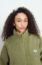 Campera de corderito con efecto shearling, color verde oliva, de corte holgado, cuello alto y cierre frontal completo. Presenta un pequeño parche con logo en el pecho.