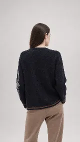 Sweater de lana color beige con calce holgado y cuello redondo. Presenta un bordado negro con diseño de arabescos en una de las mangas y detalles de rayas y puntos en contraste en el ruedo y los puños.