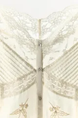 Top de algodón color crema con escote redondo, tirantes anchos y cierre frontal con botones. Presenta detalles de plisados, encaje combinado, bordados florales y aplicaciones de abalorios, con el bajo acabado en ondas.