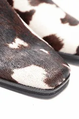Bota corta de cuero con pelo de vaca en diseño animal print de manchas marrones y blancas, con punta cuadrada, taco bajo y correa de cuero negro con tachas y una argolla metálica alrededor del tobillo.