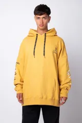 Buzo canguro amarillo ocre oversize de algodón rústico con capucha, cordones negros con letras blancas, bolsillo frontal y puños y cintura acanalados. Presenta estampados en el frente y las mangas.