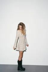 Vestido corto de algodón con estampado de cuadros en tonos beige y marrón. Presenta escote en pico, manga larga con corte evasé y bajo con volantes. Incluye cierre frontal con ganchos metálicos y cremallera lateral oculta.