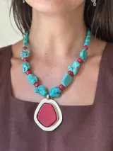 Collar corto con cuentas irregulares de magnesita turquesa y cuentas redondas de acrílico rojo, intercaladas. Incluye un dije central grande de forma irregular con borde plateado y piedra roja lisa en el centro.