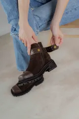 Bota corta de gamuza marrón con puntera de cuero liso, suela track negra y correa desmontable con tachas metálicas.